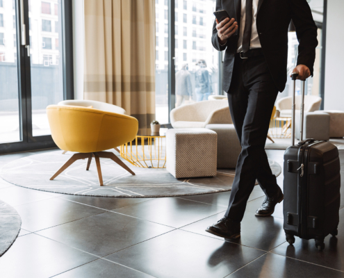 Cropped photo of successful businessman wearing suit holding smartphone and walking with suitcase in hotel lobby